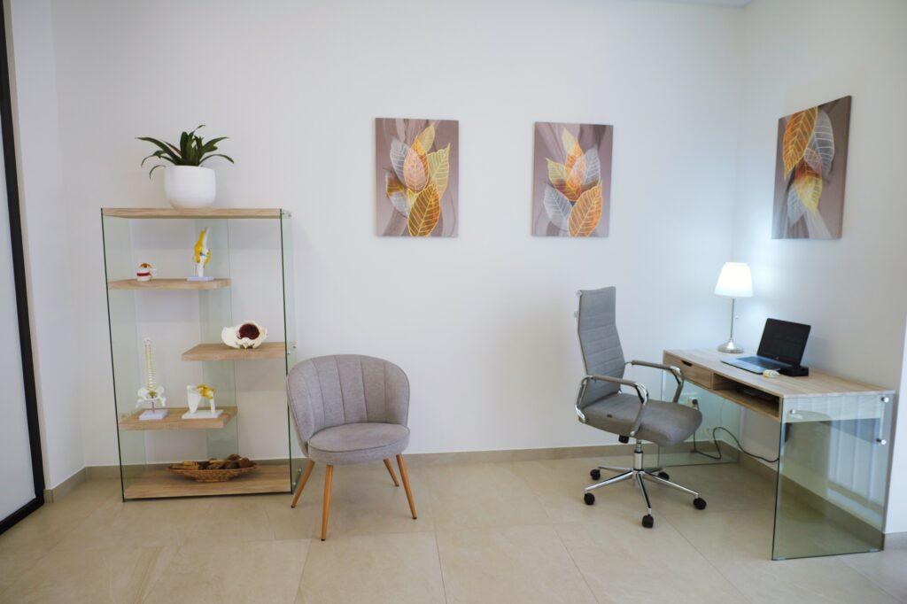 Despacho de consulta con escritorio y una estantería de cristal con modelos anatómicos de la columna y articulaciones.